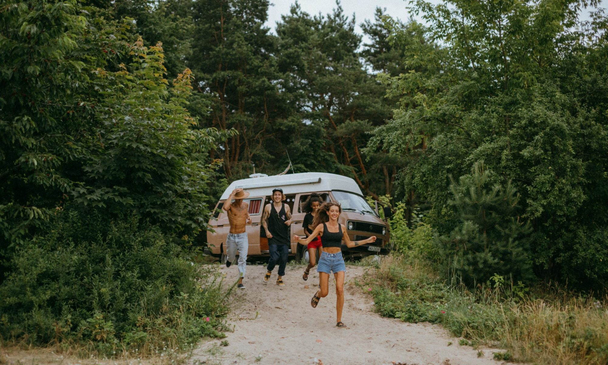 Group of friends running joyfully from a camper van in a lush forest setting during summer.